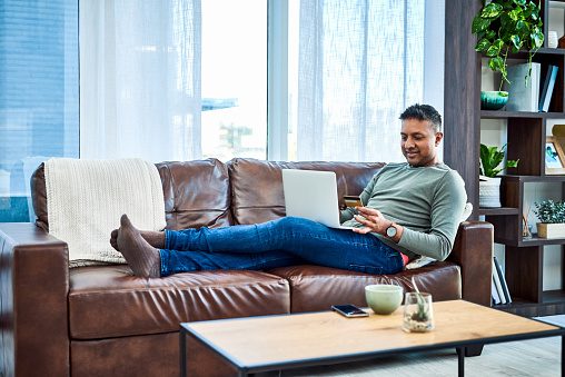 Shot of a man using a laptop and credit card on the sofa at home