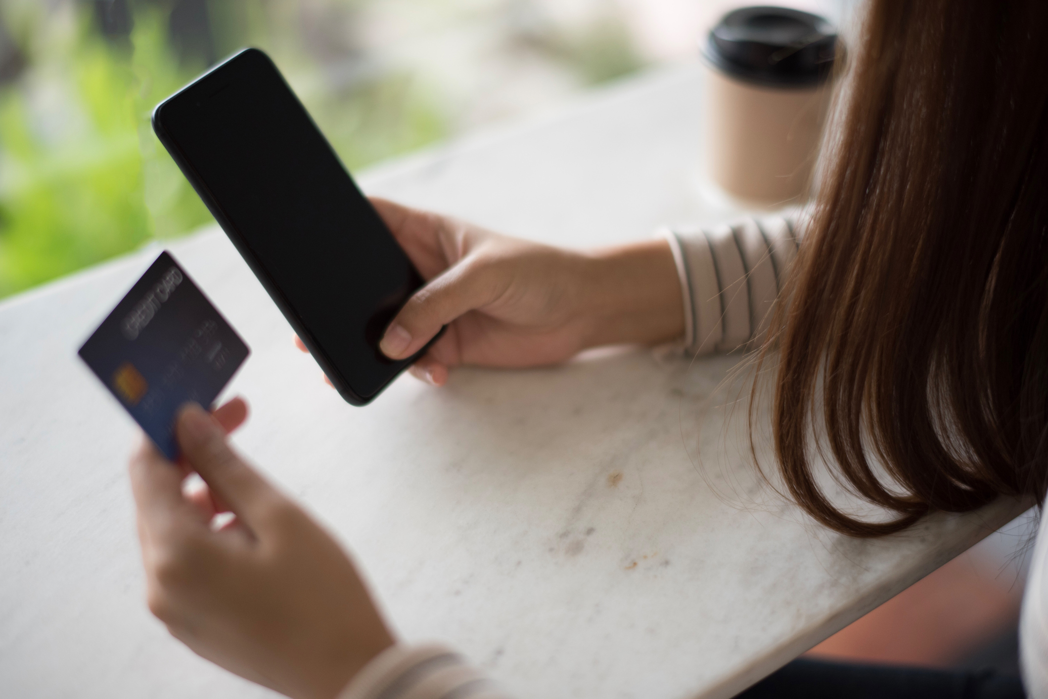 Closed up and focus on hand of young woman using a smartphone and other hand hold a credit card which is shopping online and sitting on table near big mirror at cafe. online payment concept.